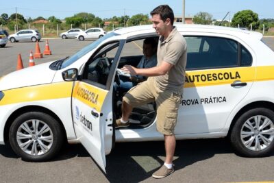 Candidato de bermuda entra no carro da autoescola para a prova prática do Detran, iniciando exame de direção.