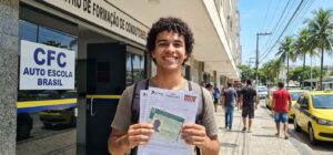 Jovem brasileiro de 18 anos, sorrindo, segurando formulários da CNH em frente a um centro de formação de condutores.