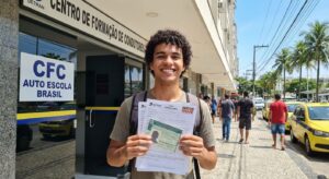 Jovem brasileiro de 18 anos, sorrindo, segurando formulários da CNH em frente a um centro de formação de condutores.