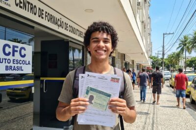 Jovem brasileiro de 18 anos, sorrindo, segurando formulários da CNH em frente a um centro de formação de condutores.